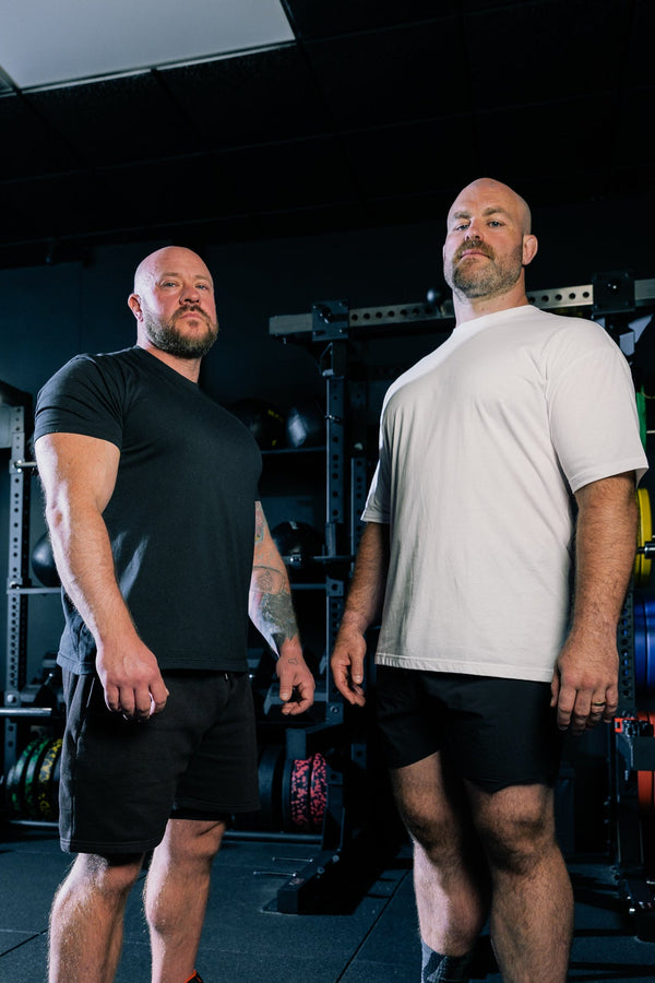 Two men standing in a dark gym environment, one wearing a black t-shirt and shorts with white sneakers, and the other wearing a white t-shirt and black shorts.